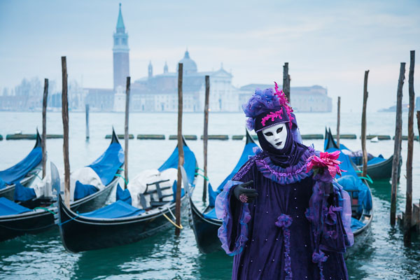 Photo of a purple costumed and masked woman standing in front of some gondolas with San Giorgio Maggiore in the background and some fog during the carnival in Venice, Italy