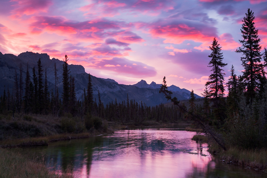 Photo of a glorious sunrise with dramatic pink clouds over Glory Hole, the pink reflecting off the water of a small river, trees and the Canadian Rocky Mountains, Jasper National Park, Alberta, Canada
