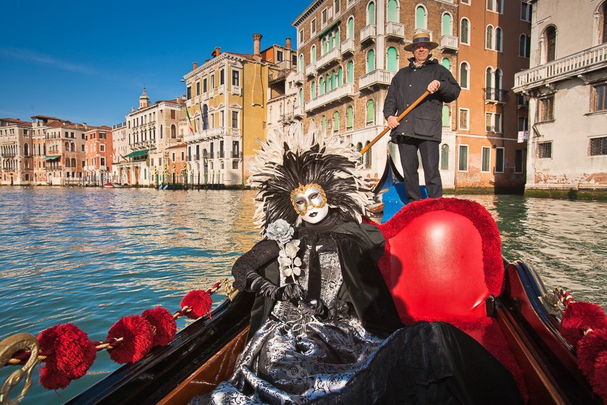 Photo of a costumed and masked woman in a gondola on the Canale Grande in Venice, Italy, with historic houses in the background